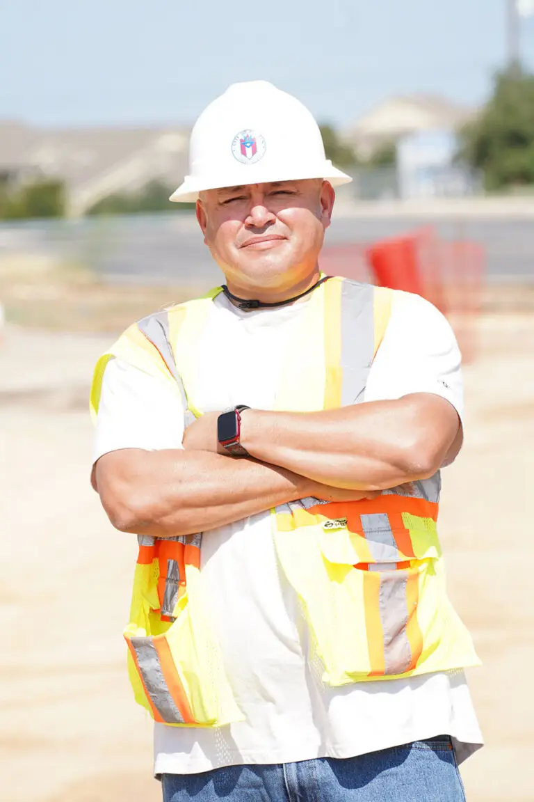 A man in a white hard hat and yellow safety vest stands with arms crossed at a construction site