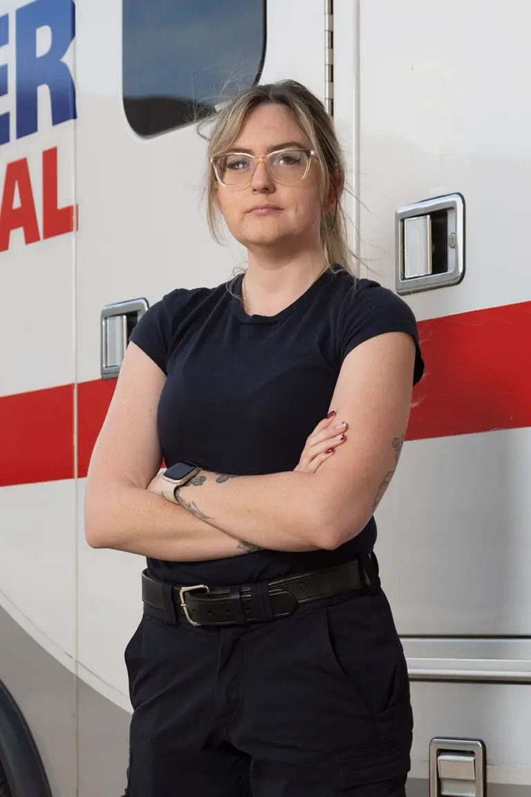 A woman with blonde hair and glasses stands with arms crossed in front of an ambulance