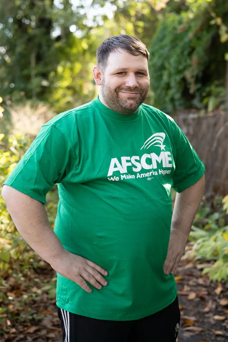 A man in a green AFSCME 'We Make America Happen' t-shirt stands smiling in an outdoor garden setting