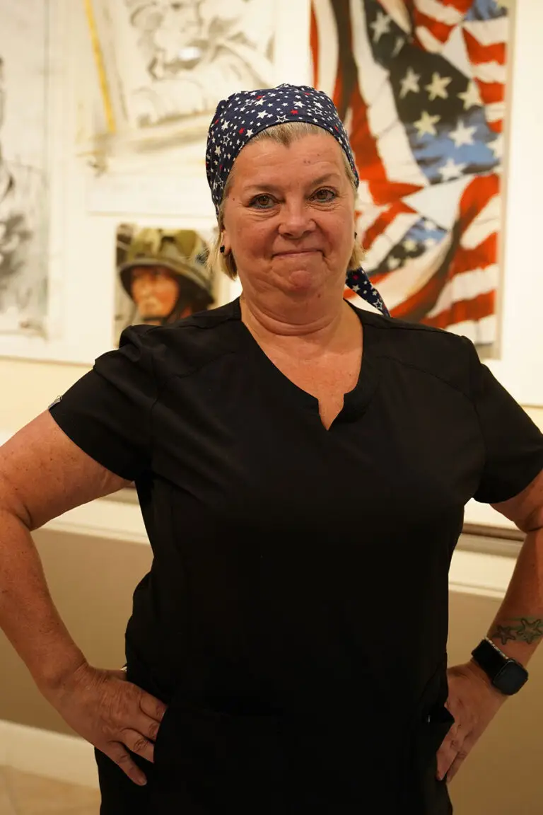 A woman in a black shirt and a star-patterned bandana stands with hands on hips in front of artwork featuring an American flag