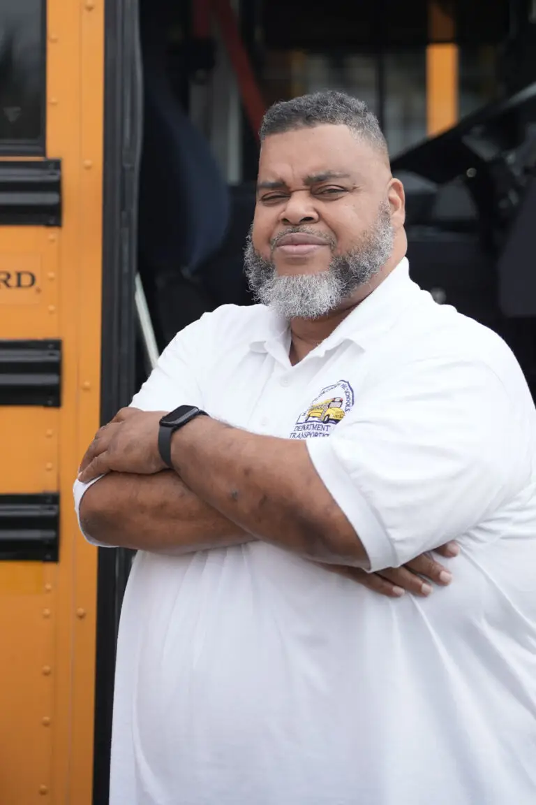 A man in a white polo shirt stands with arms crossed in front of a yellow school bus