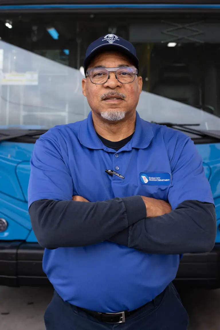 A man in a blue Kansas City Aviation Department polo shirt and baseball cap stands with arms crossed in front of a blue bus